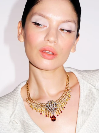 Close-up portrait of model wearing an ornate gold gemstone necklace and white jacket against a neutral background