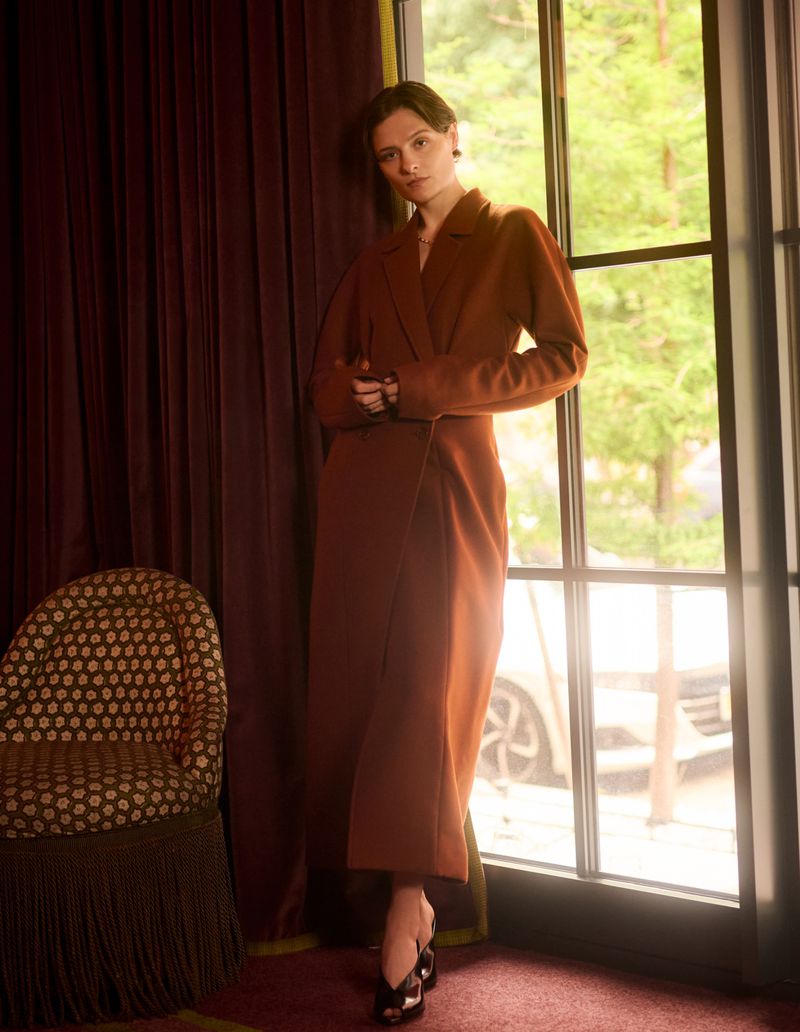 Louisa Jacobson wearing a long rust brown tailored coat standing by a sunlit window next to a patterned fringed armchair.