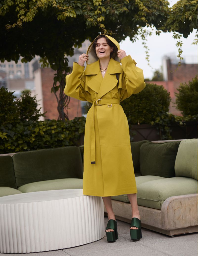 A woman poses on a rooftop in a yellow raincoat, her hands on the hood, against a backdrop of trees and buildings under an overcast sky.