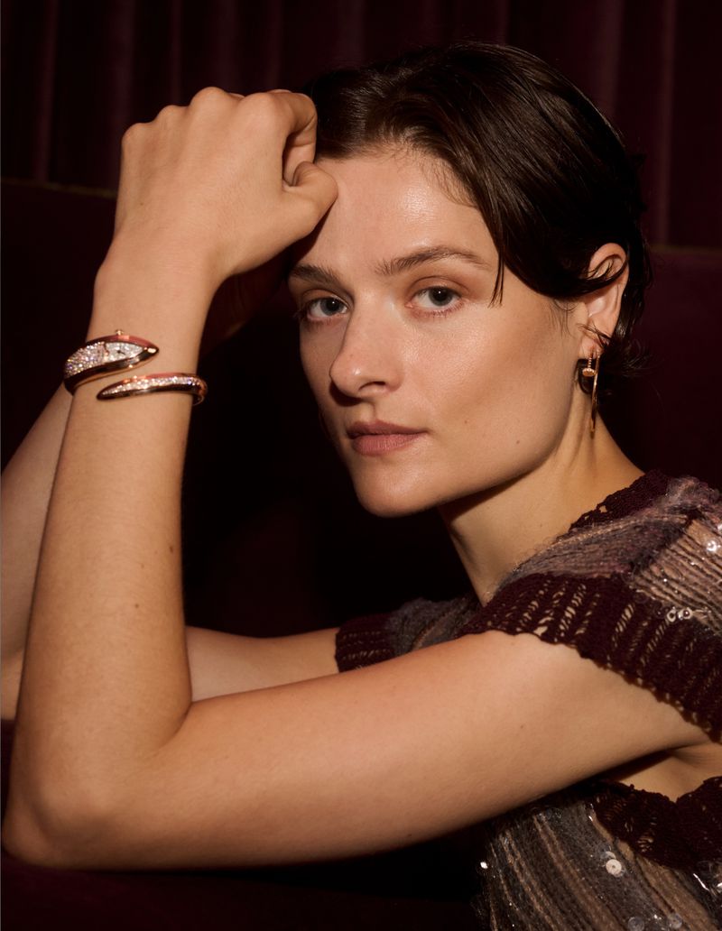 Brunette woman poses indoors wearing a sequined top and gold bracelet with diamonds in a medium shot against a dark backdrop.
