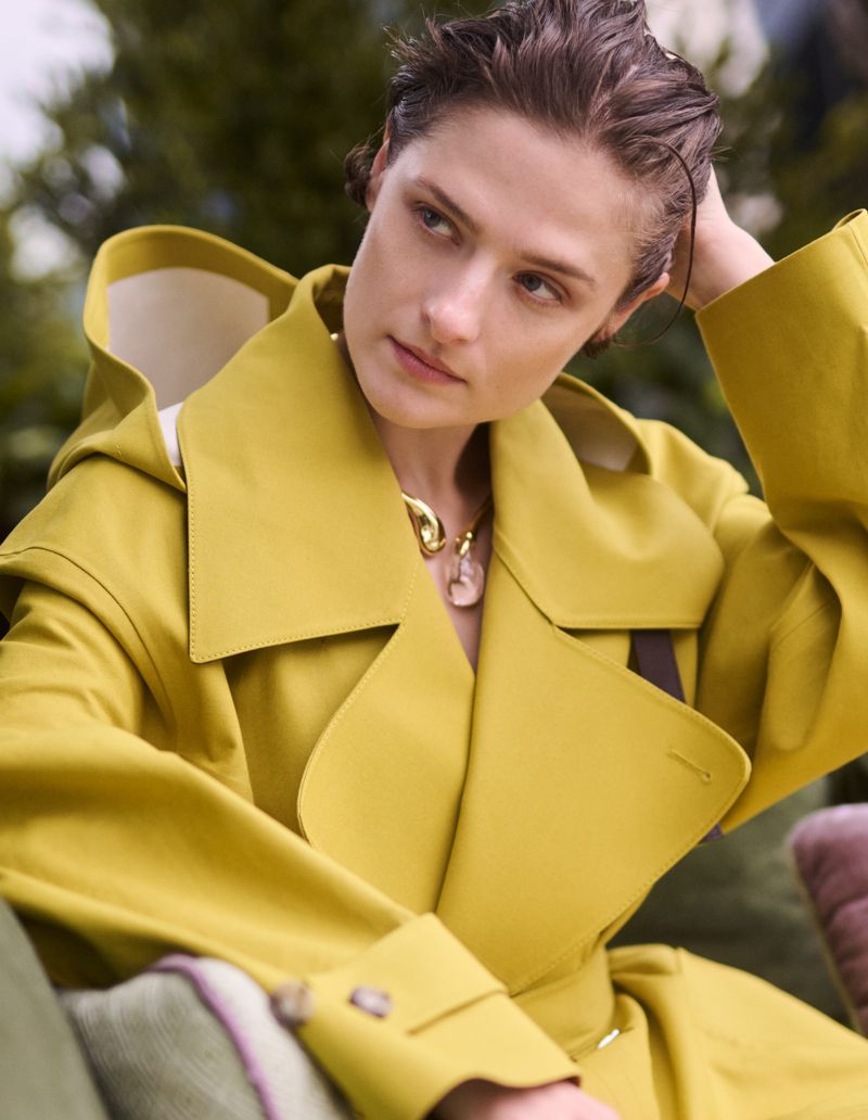 Woman in a yellow trench coat poses outdoors with her hand in her hair and a gold necklace, soft light.
