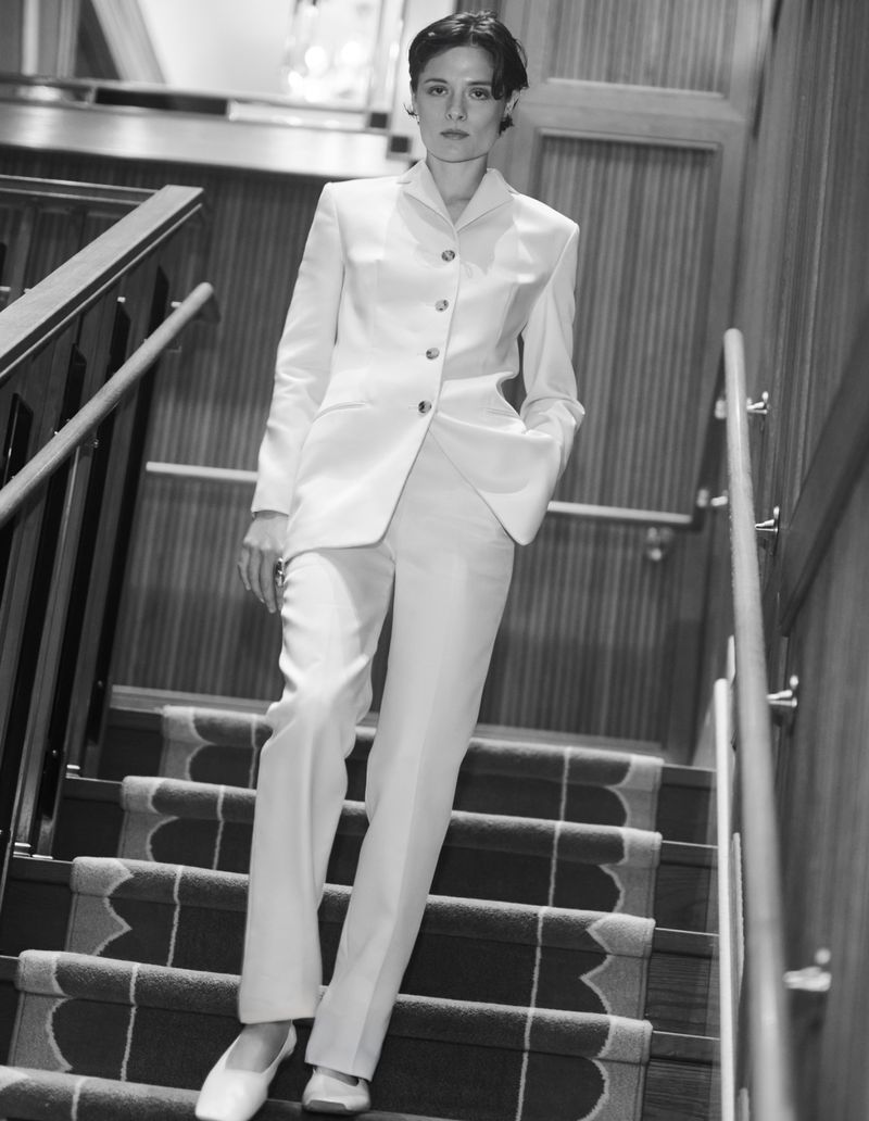 Monochrome full-body portrait of an adult woman in a light-colored suit walking down carpeted stairs in an indoor setting.