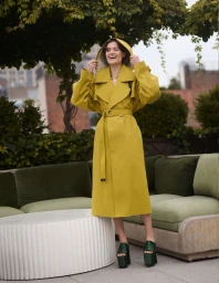Louisa Jacobson laughing in a hooded yellow trench coat and dark green platform heels on an outdoor terrace for Vestal Magazine.