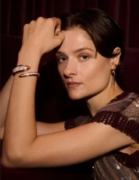 Louisa Jacobson posing with her hand raised, wearing a diamond serpent cuff bracelet and a sequined knit top against a dark backdrop.