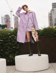 Model in a lavender coat and skirt standing on a round white pedestal with city buildings and construction cranes in the background