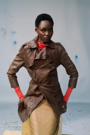 Model Caren Jepkemei in a brown leather trench coat and red top against a grey seamless studio backdrop.