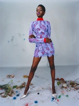 Model Caren Jepkemei posing in a patterned purple mini dress and white heels against a weathered studio background