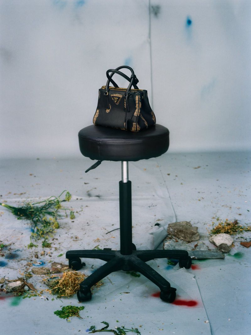 Black leather handbag resting on a round rolling stool against a weathered studio wall.