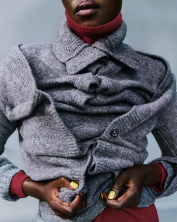 Model Caren Jepkemei wearing a layered grey knit cardigan over a red turtleneck sweater against a grey background.