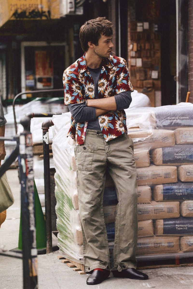 A man wearing a patterned short-sleeve shirt and khaki trousers leaning against stacked bags outdoors on a city sidewalk.