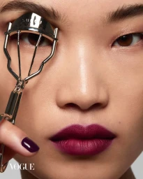 Close-up portrait of model holding a metal eyelash curler near her eye, wearing dark plum lipstick, for Vogue Thailand Beauty.