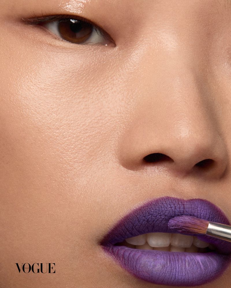 Close-up portrait of a model having purple lipstick applied with a brush for Vogue Thailand.