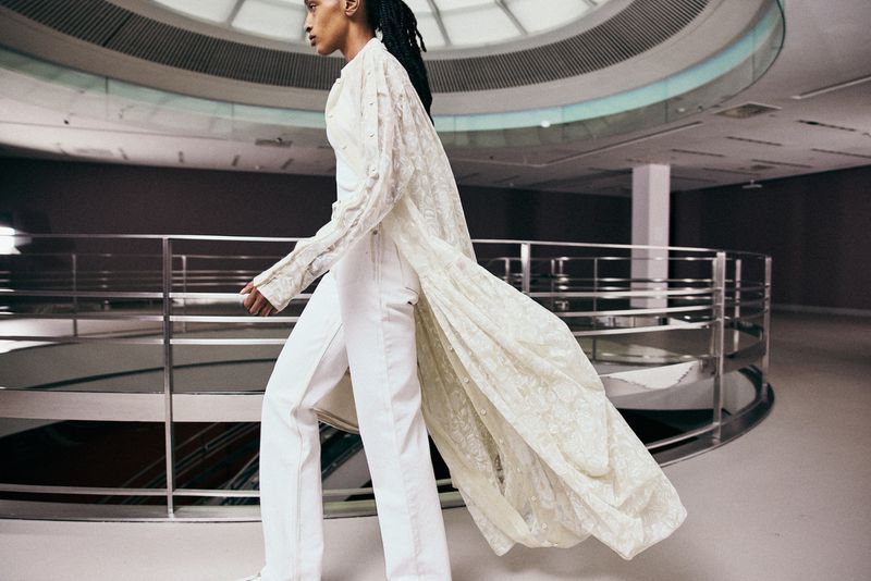 Model walking in a long cream lace coat and white trousers inside a modern multi-level building with a circular atrium and metal railings.