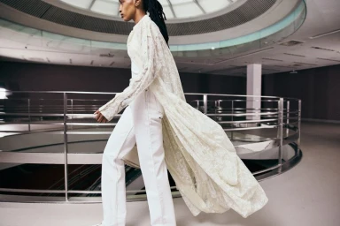 Model walking in a long cream lace coat and white trousers inside a modern multi-level building with a circular atrium and metal railings.