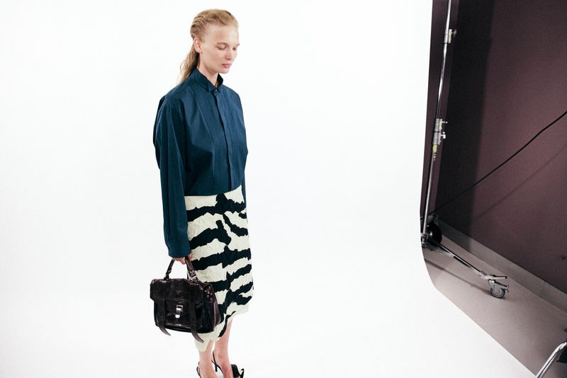Model in a teal button-down shirt and patterned skirt holding a dark handbag in a white studio setting.