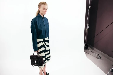 Model in a teal button-down shirt and patterned skirt holding a dark handbag in a white studio setting.