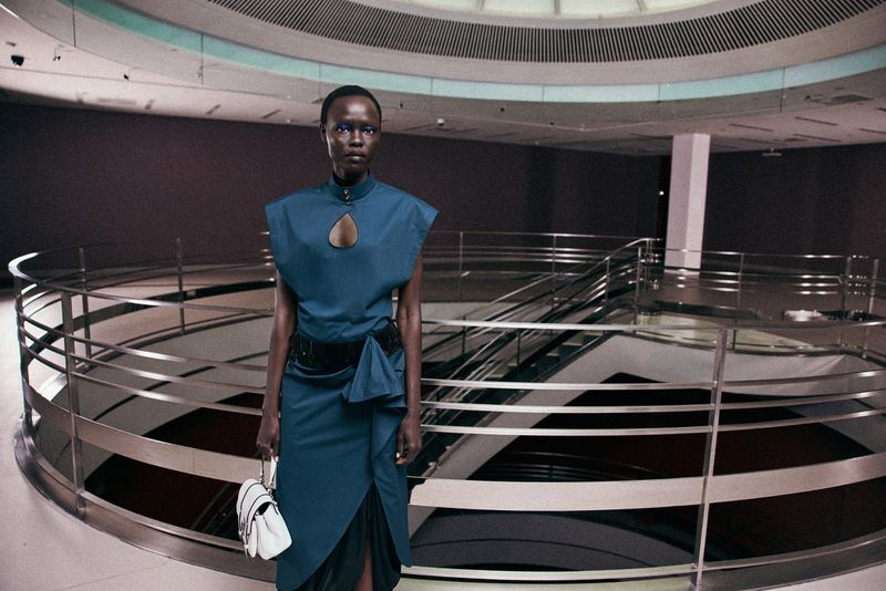 Model wearing a dark teal sleeveless dress and holding a white handbag standing on a balcony overlooking a modern interior atrium.