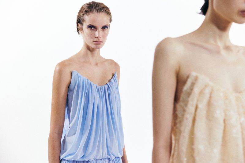 Model in a light blue gathered strapless dress against a bright white background with another person partially visible in the foreground