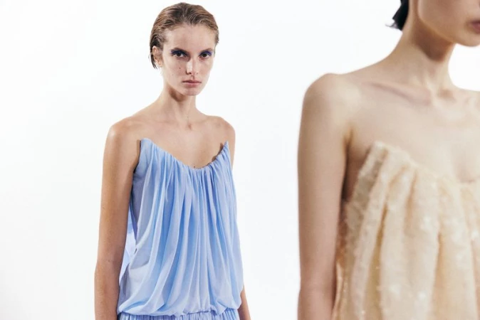 Model in a light blue gathered strapless dress against a bright white background with another person partially visible in the foreground