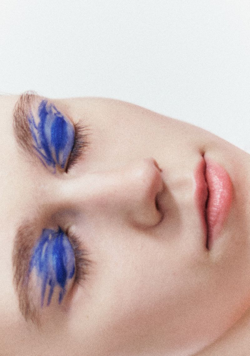 Close-up portrait of a model with vivid blue painted eyeshadow on closed eyelids against a neutral background.