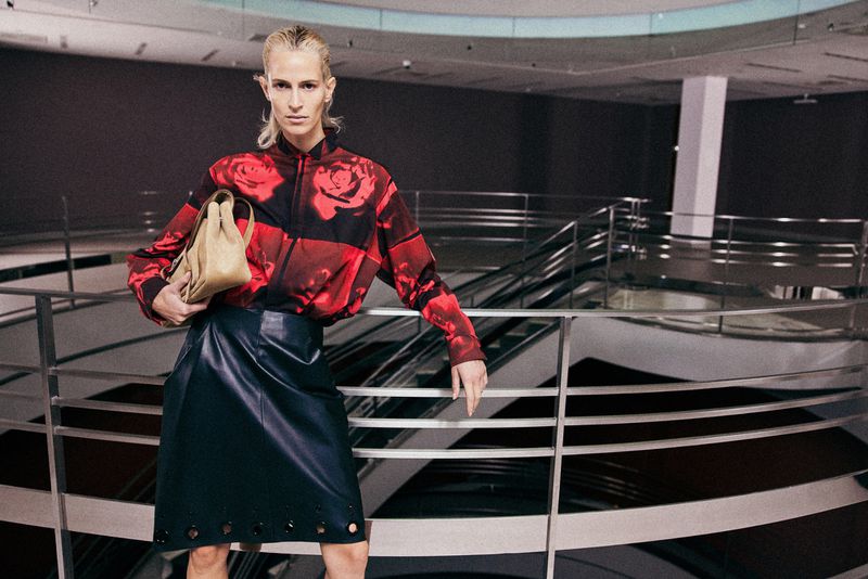 Model in red rose print shirt and dark skirt holding a tan handbag, leaning on a metal railing in an interior space.