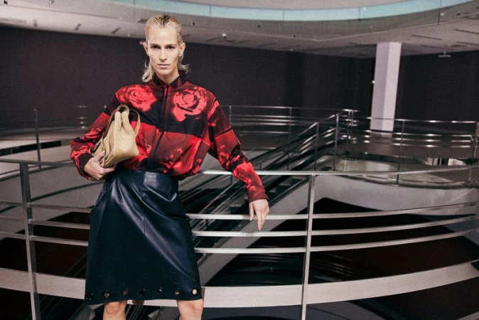 Model in red rose print shirt and dark skirt holding a tan handbag, leaning on a metal railing in an interior space.