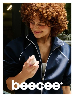 Woman with curly red hair wearing a dark blue zip-up jacket holding a small spray bottle for the Beecee 2026 campaign.