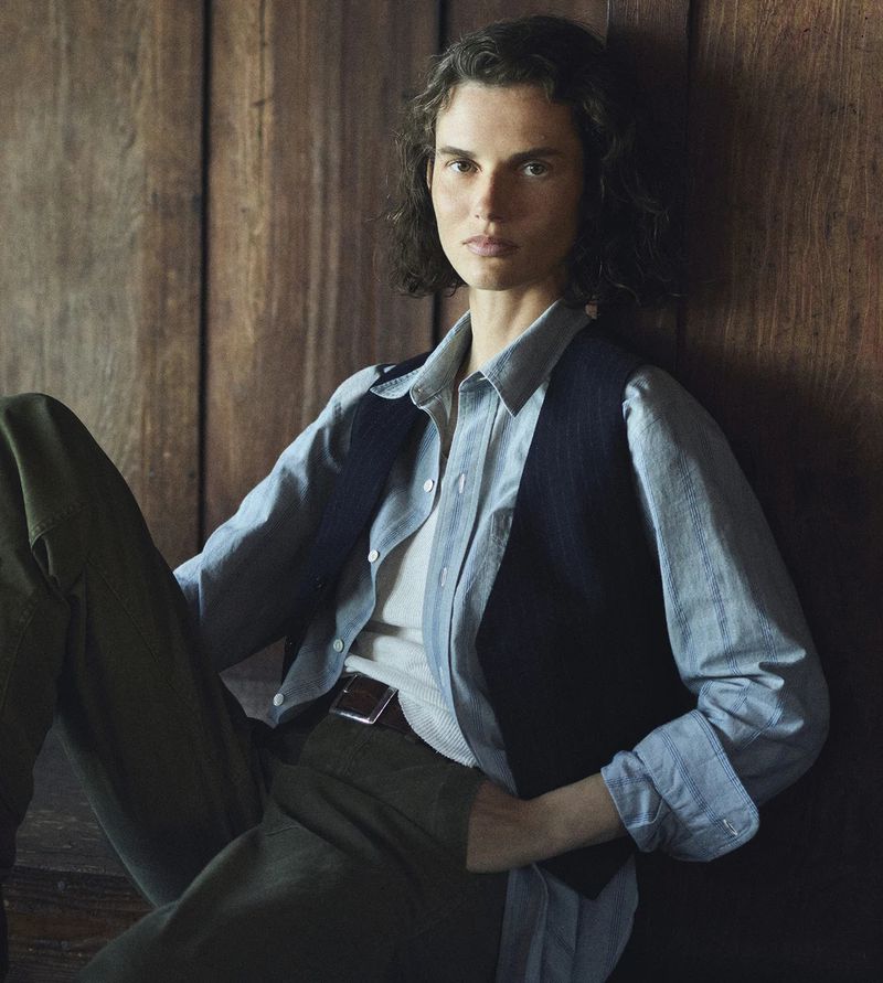 Model Giedre Dukauskaite wearing a blue shirt and dark vest against a wood background for Buck Mason.