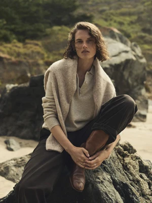 Model Giedre Dukauskaite wearing a beige sweater and dark trousers seated on rocks at the coast for Buck Mason