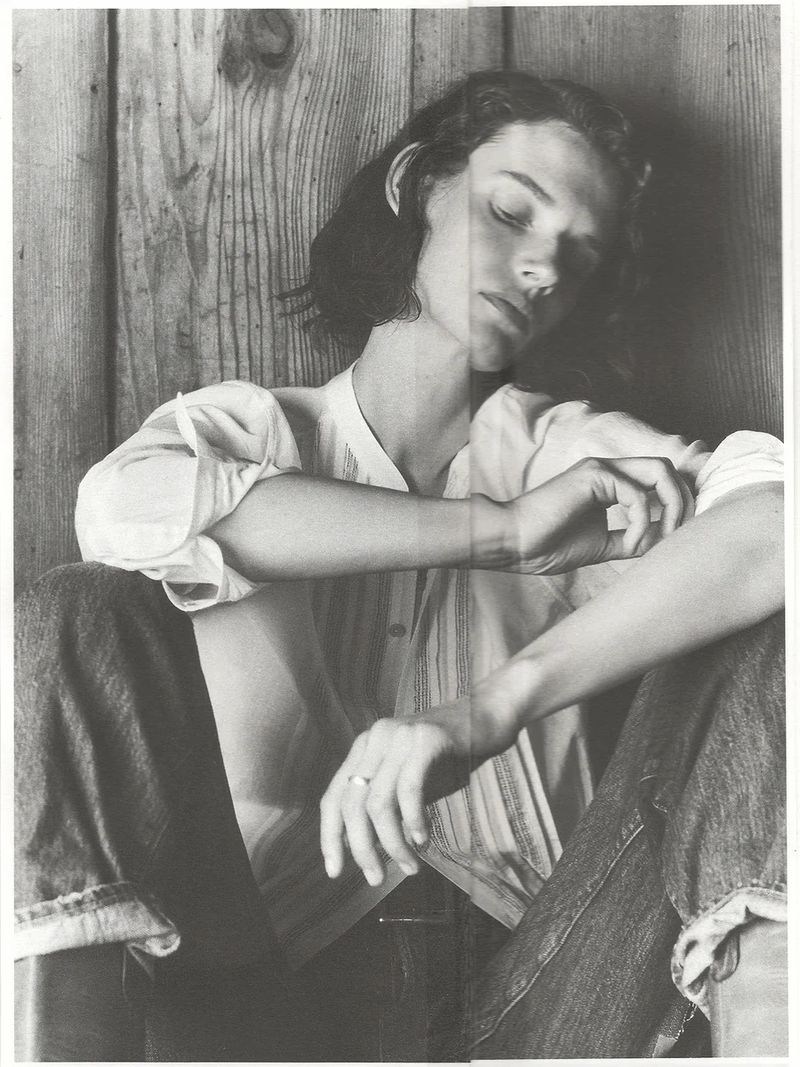 Black and white portrait of a model sitting against a wooden wall wearing a light button-down shirt and dark jeans.