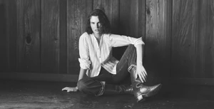Model Giedre Dukauskaite seated against a dark wood wall wearing a white button-down shirt and dark jeans