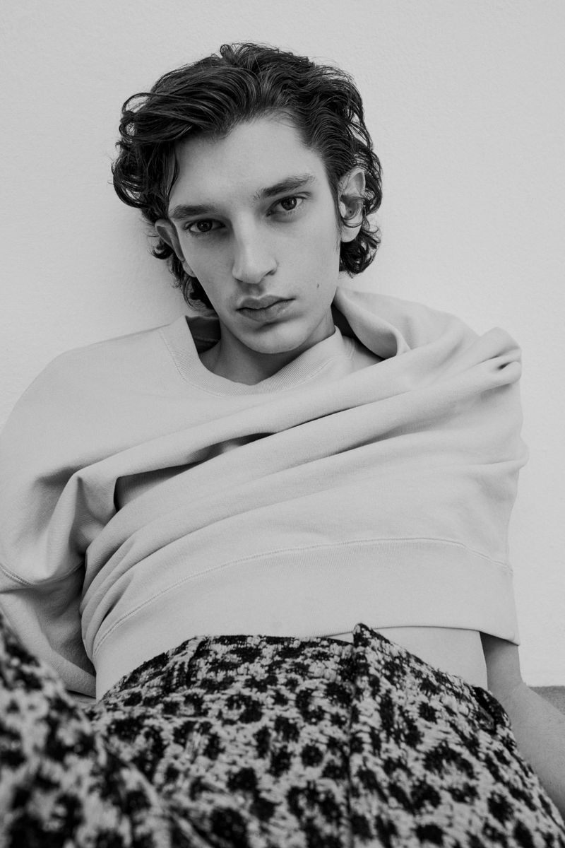 Monochrome portrait of a young adult male with curly dark hair wearing a sweatshirt and patterned pants against a plain white wall.