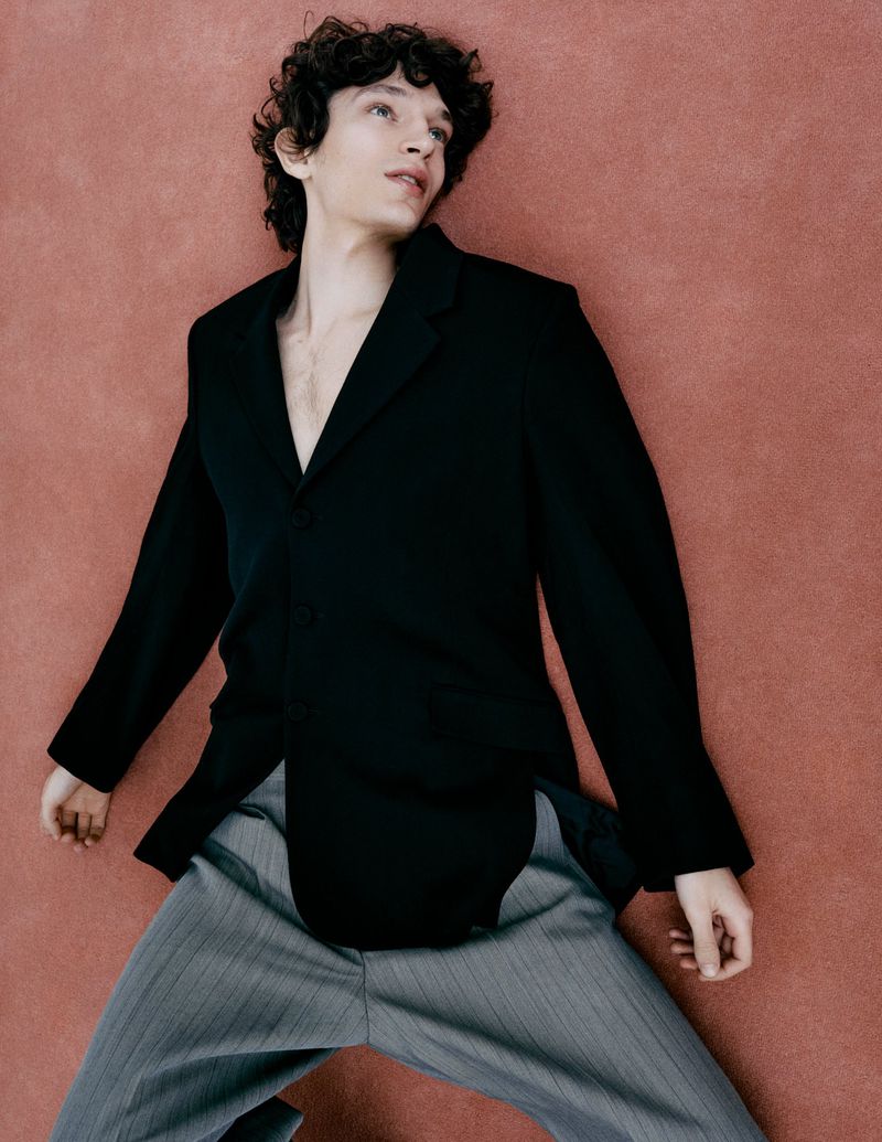 Man in a black blazer and gray pants lies against a rose-colored backdrop, fashionably posed in a studio setting with soft, diffused lighting.