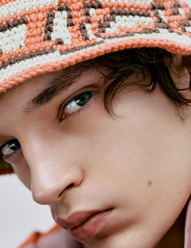Close-up portrait of a young adult male with blue eyes and light skin, wearing a patterned crochet bucket hat against a neutral background.