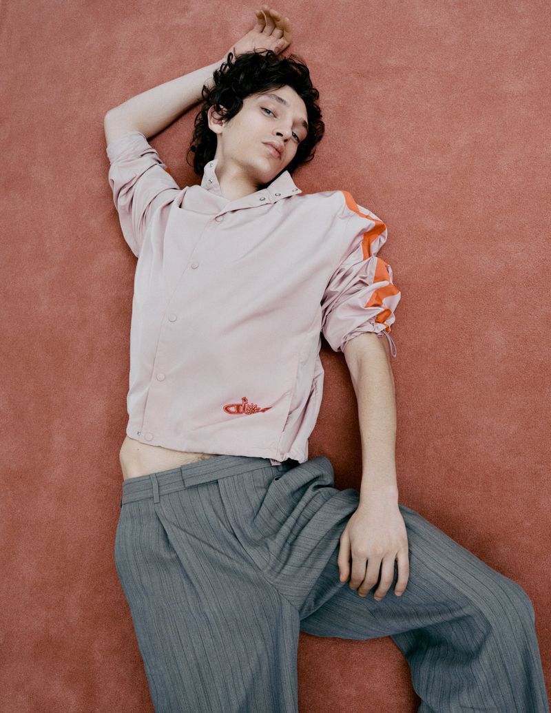A young adult male is lying on a textured pink surface in a relaxed pose, wearing a pink cropped jacket and grey striped pants in a fashion shot.