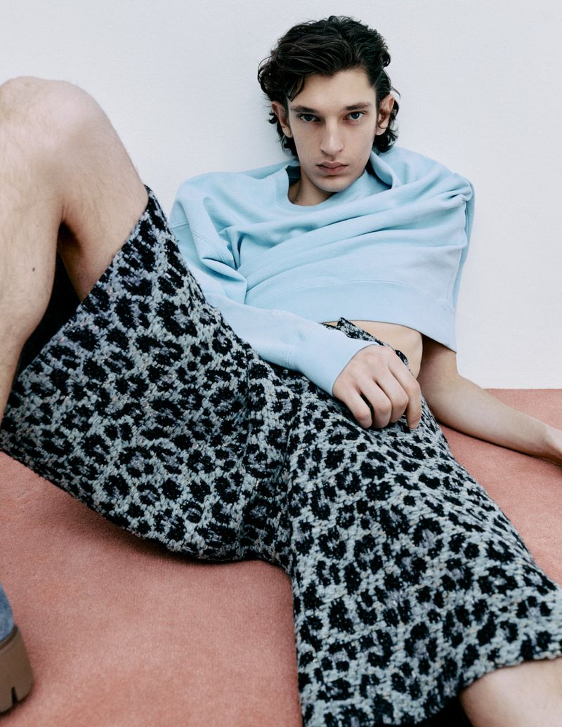 A young adult man in a light blue sweatshirt and animal print pants is reclining against a white wall with a pink baseboard.