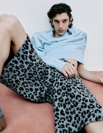 A young adult man in a light blue sweatshirt and animal print pants is reclining against a white wall with a pink baseboard.