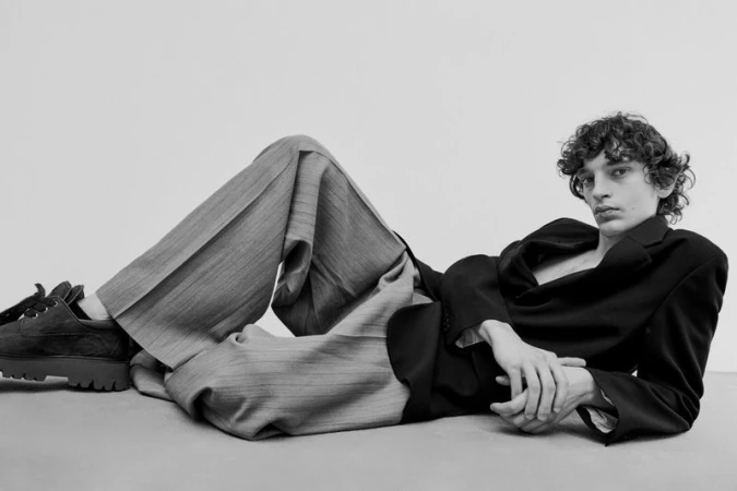 A young adult with curly hair poses in a black jacket and striped trousers while lying on a neutral surface in a studio setting.