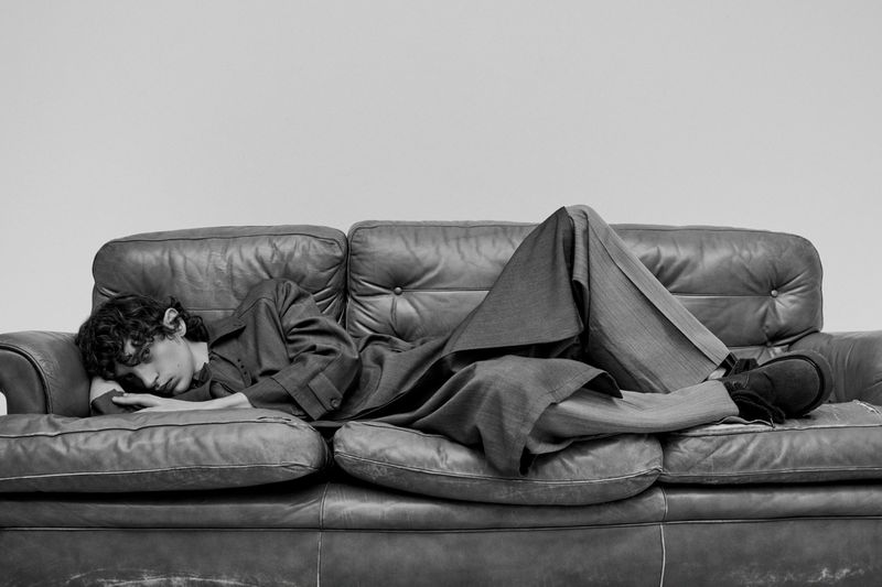 A young adult male with curly hair rests on a leather couch, wearing a matching suit in a minimalist studio setting, captured in black and white.