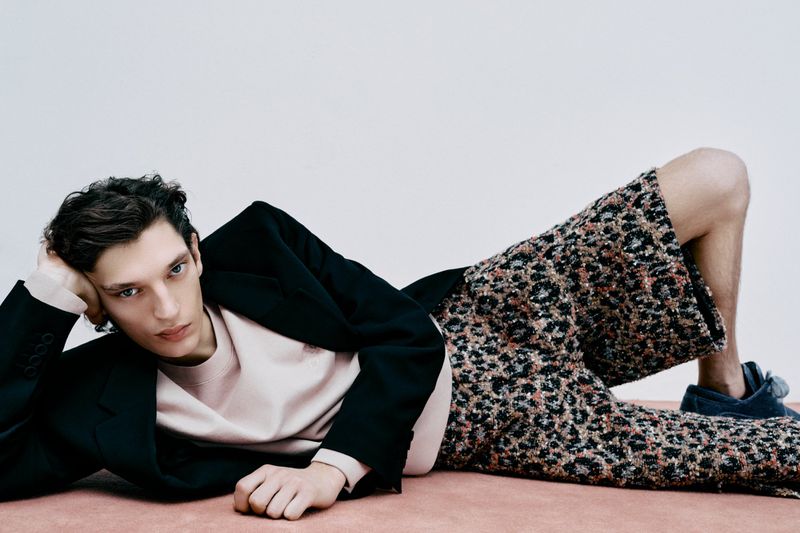 Male fashion model poses in a relaxed prone position wearing a black blazer, light pink top, and patterned culottes against a neutral backdrop.