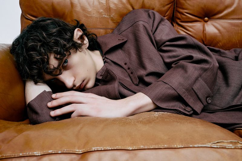 A young adult male with curly hair rests on a brown leather couch, wearing a brown button-down shirt. He has fair skin and blue eyes.