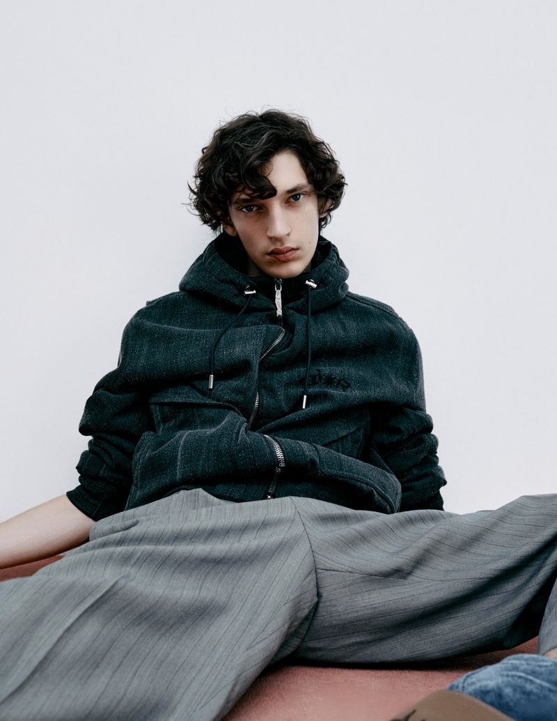 An adult male model poses indoors wearing a hooded denim jacket and grey striped pants, leaning on a mauve surface with a white wall background.