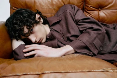 A young man with curly dark hair wearing a brown coat lies reclined on a brown leather sofa.