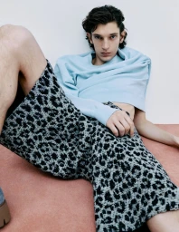 Model Cirillo wearing a light blue cropped sweatshirt and leopard print shorts leaning against a white studio wall on a peach floor.