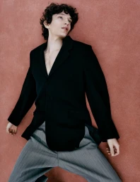 Model with dark curly hair wearing a black blazer and grey trousers against a textured red background.
