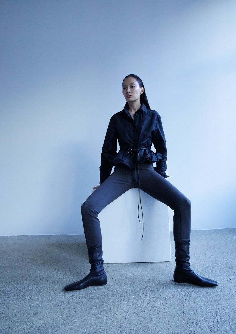 Model seated on a white block wearing a dark long-sleeve belted top and dark leggings against a grey studio wall.