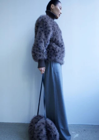 Model wearing a textured brown furry jacket and wide grey trousers holding a matching handbag against a plain background