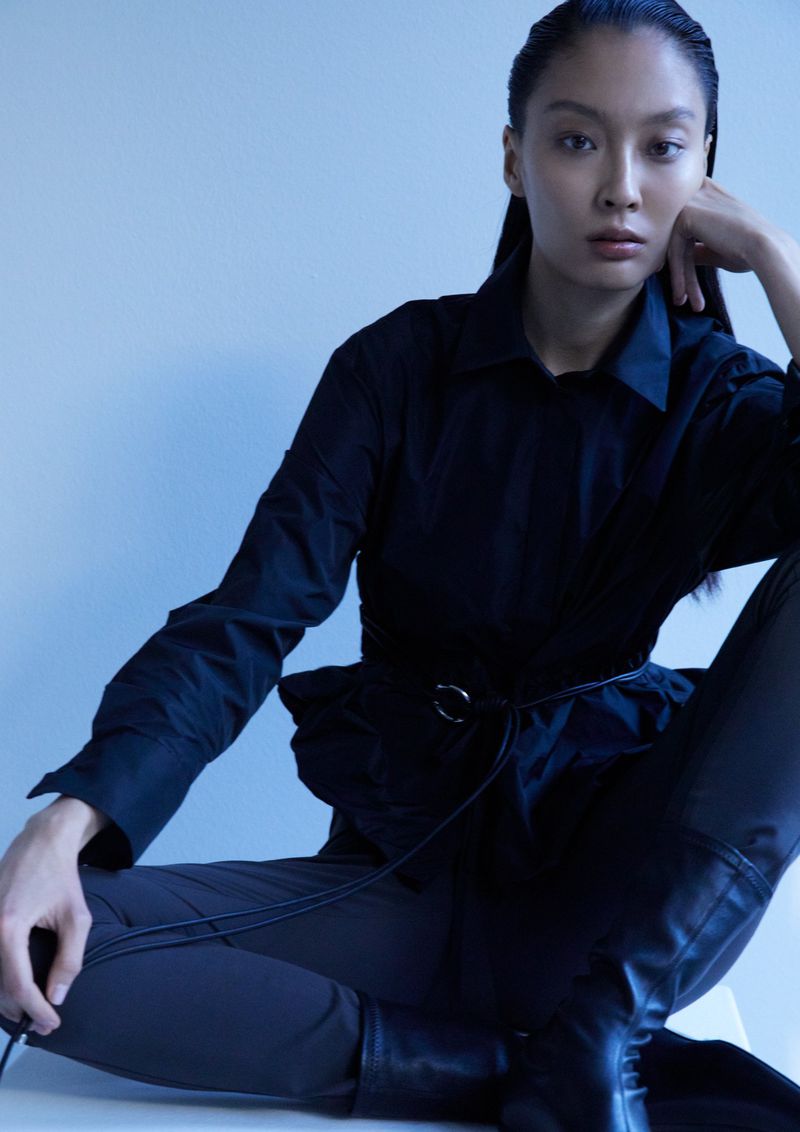 Model in a black shirt and dark trousers seated against a pale blue background for Uli Schneider FW 2026
