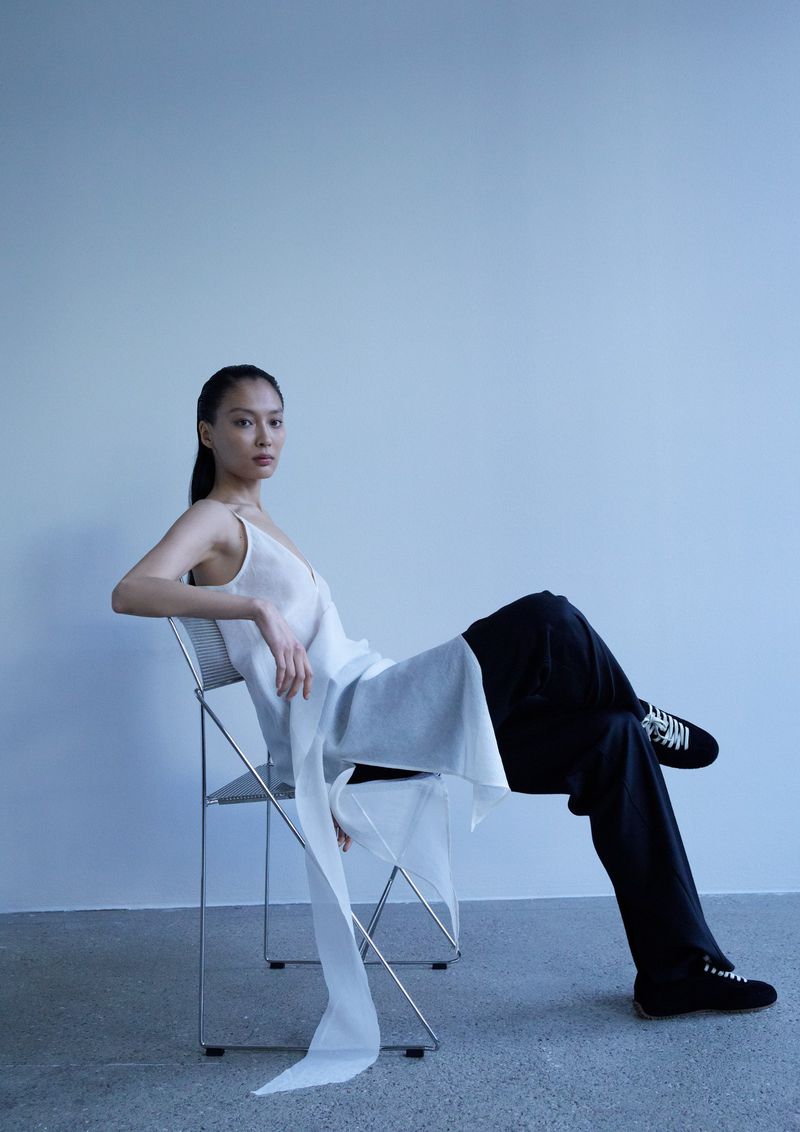Model seated on a metal wire chair wearing a white tunic top and black trousers against a neutral wall.