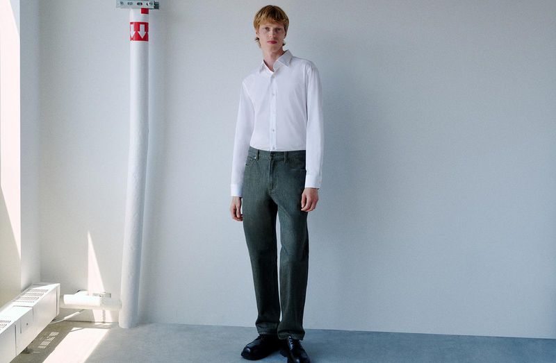 Full-length portrait of a male model wearing a white shirt and dark trousers against a plain wall.
